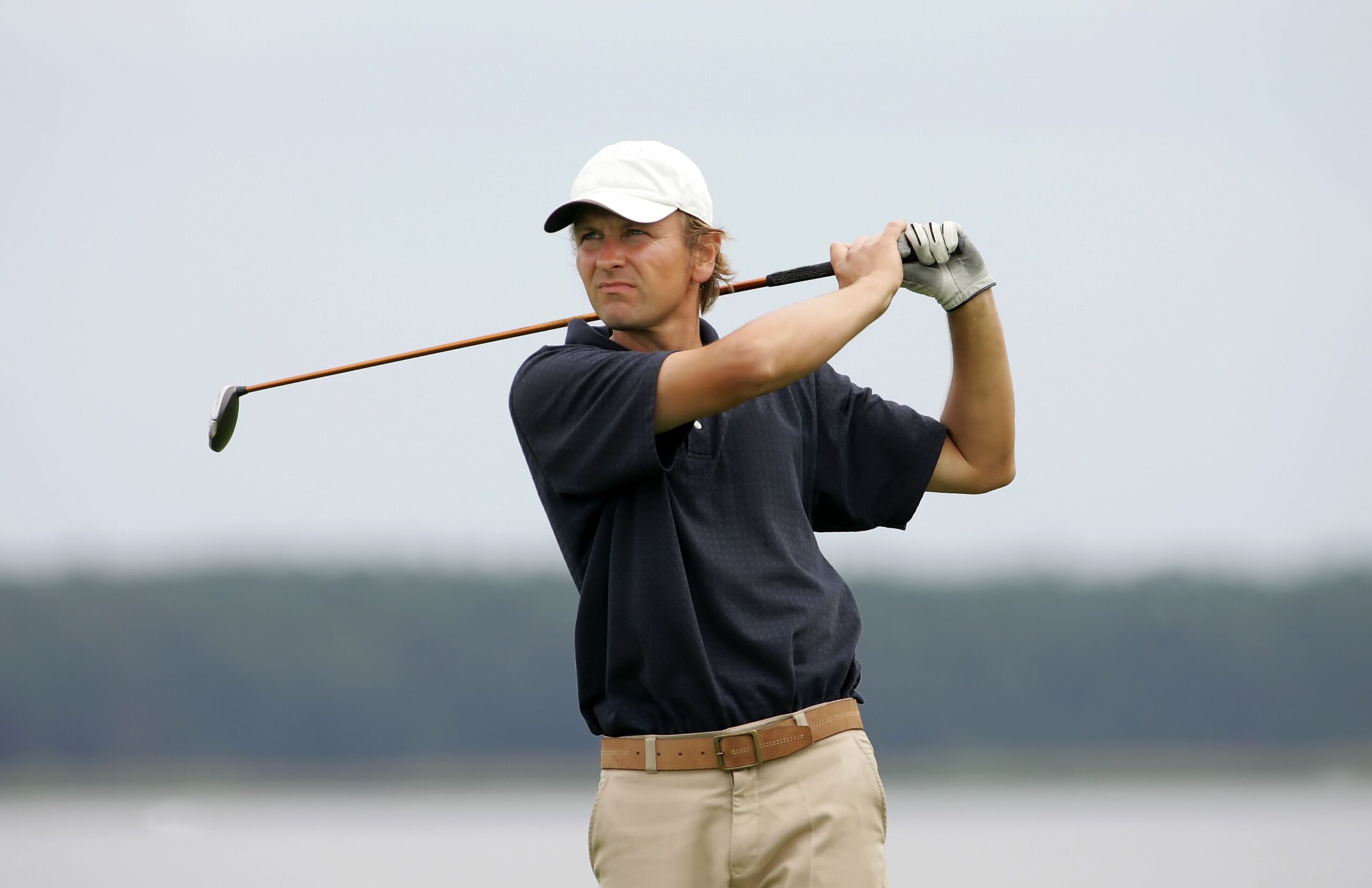 A golfer in a navy polo and white cap prepares to swing his club on a calm, overcast day, emphasizing the focus and technique of the sport.
