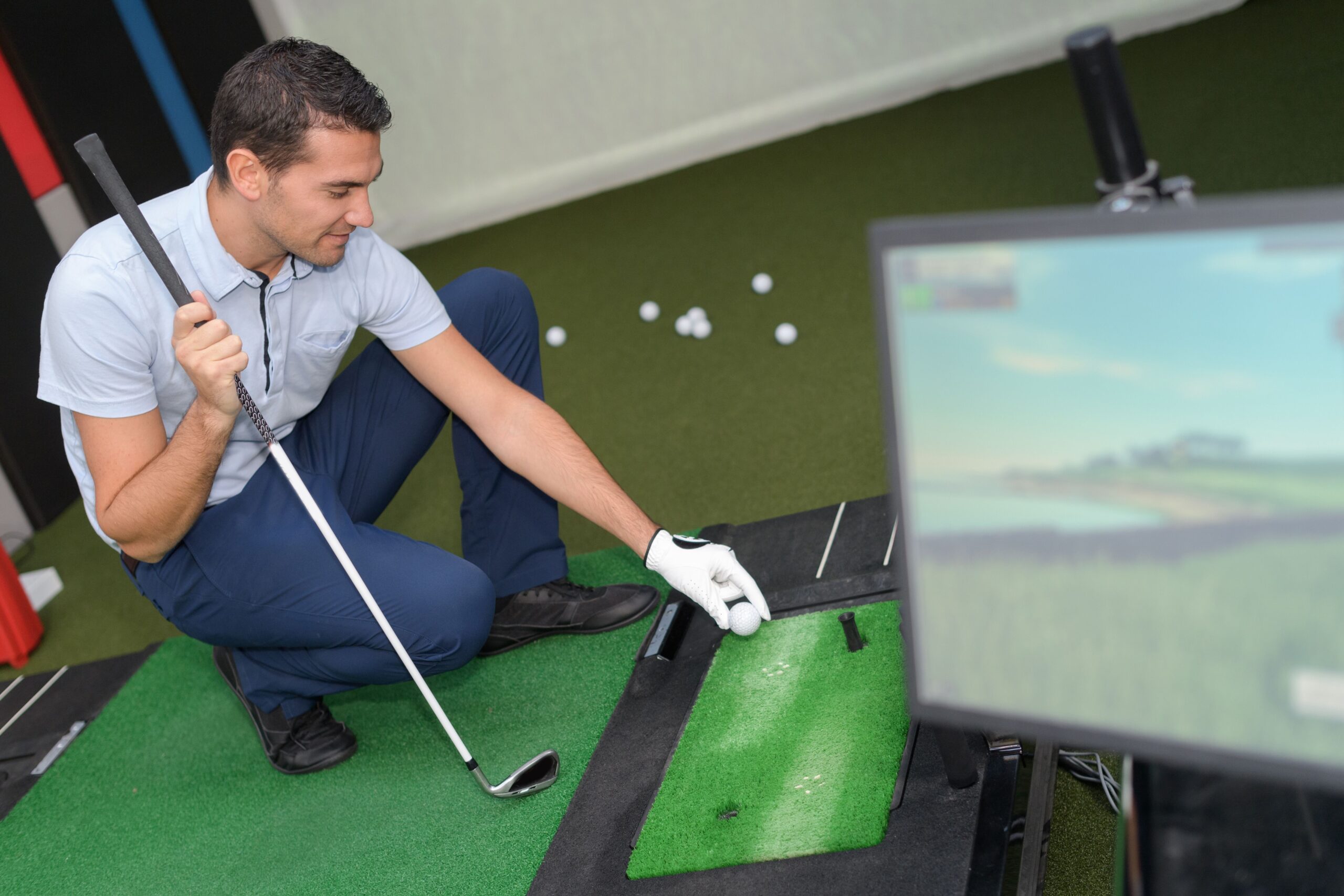 A golfer kneels to adjust a golf ball on a green mat, while a monitor displays a digital golf course, illustrating a practice or training session.
