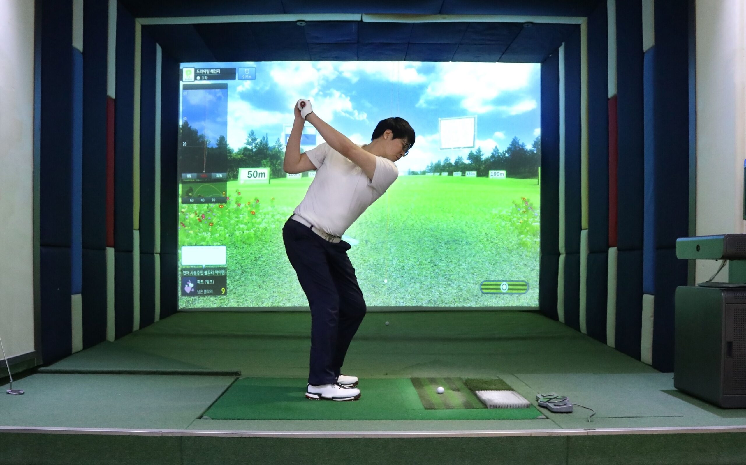 A golfer prepares to swing in a virtual golf simulator, with a realistic outdoor scene displayed on the screen, showing distance markers.