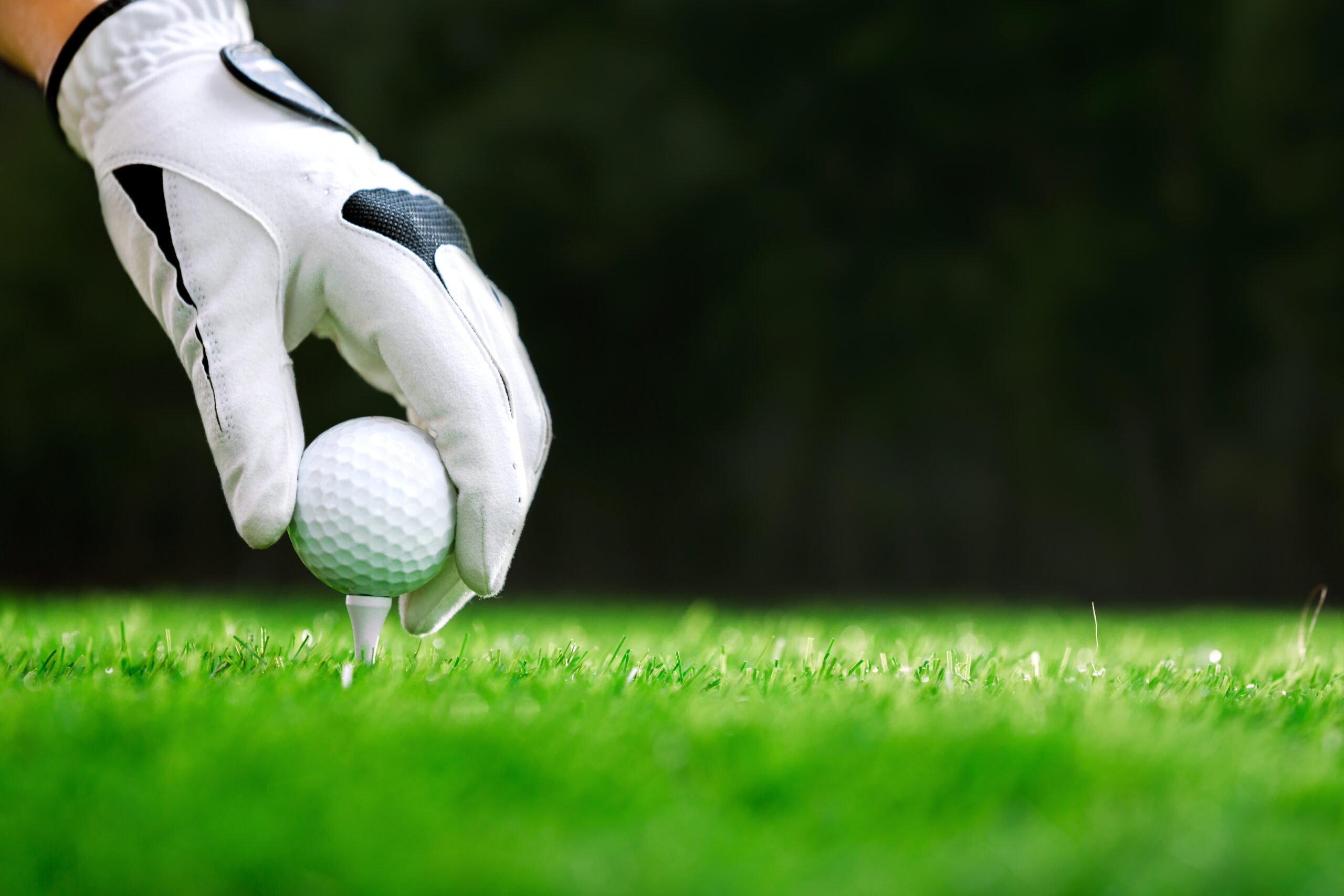 A hand in a golf glove is placing a white golf ball on a tee in lush green grass, illustrating the preparation for a golf swing.