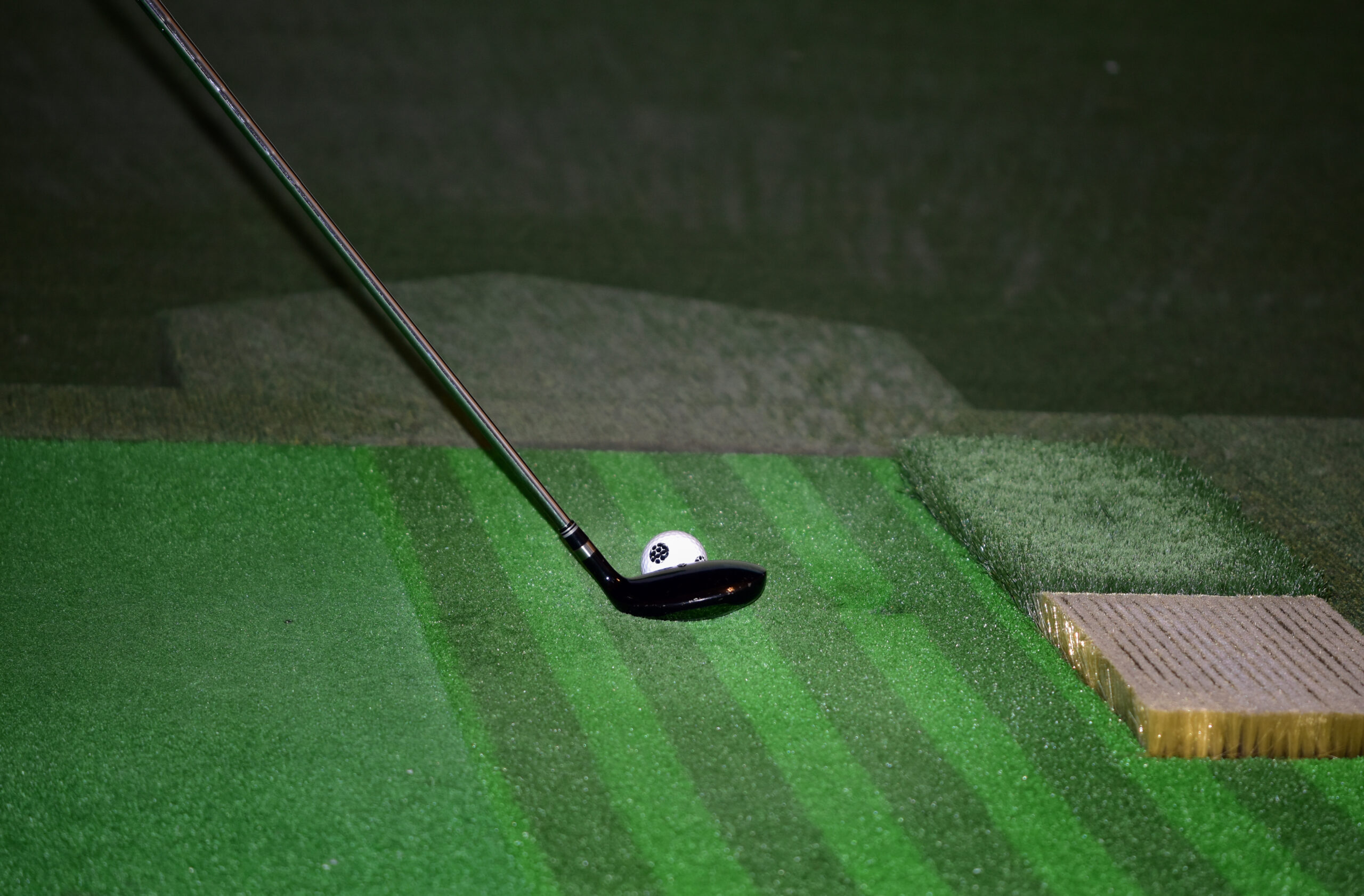 Golf club poised over a white ball on a textured green mat, illustrating a practice swing for golfers improving their technique.